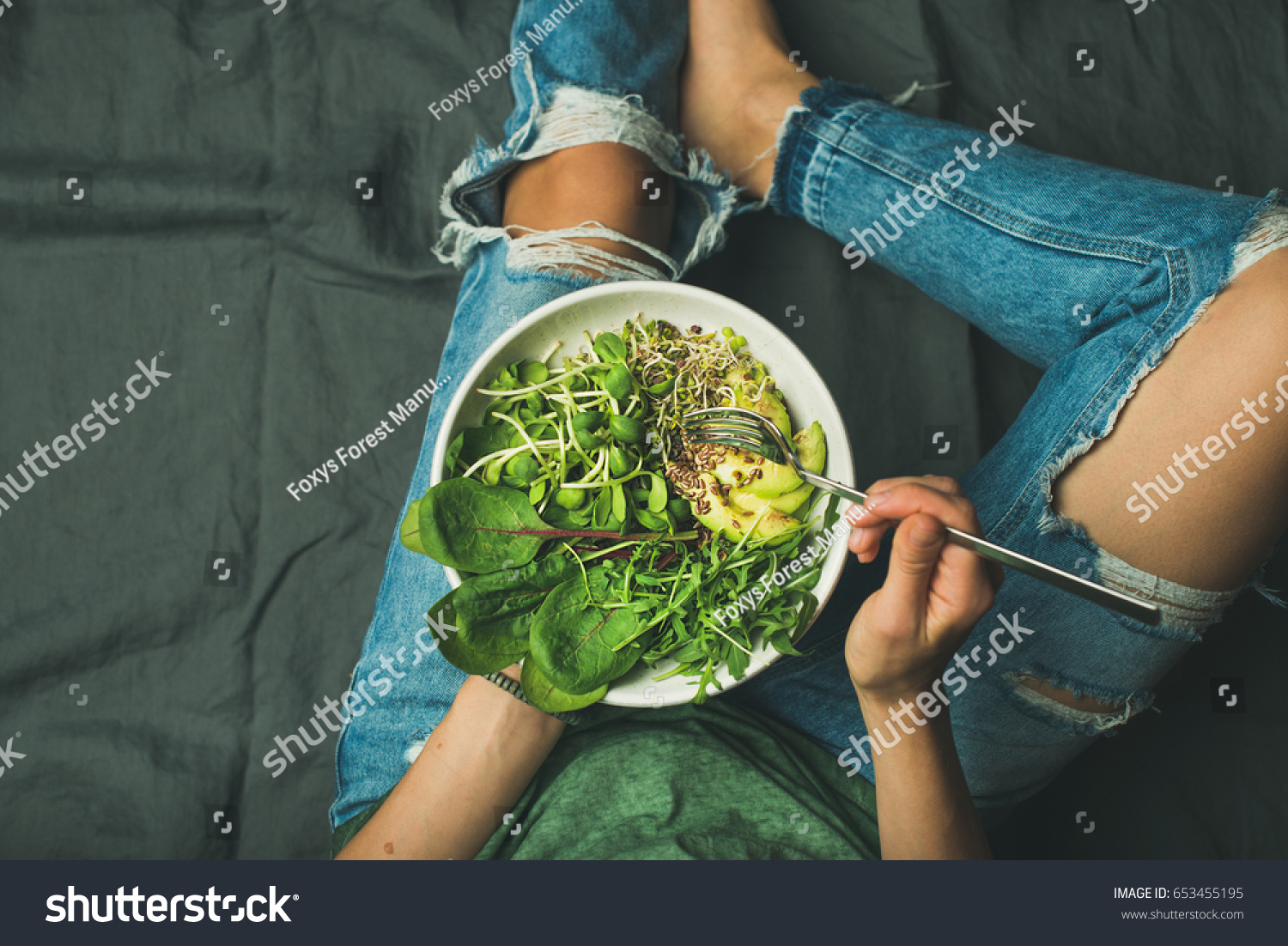 Green vegan breakfast meal in bowl with spinach, arugula, avocado, seeds and sprouts. Girl in jeans holding fork with knees and hands visible, top view, copy space. Clean eating, vegan food concept #653455195