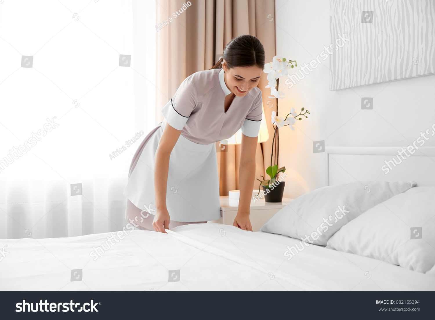 Young maid making bed in light hotel room #682155394