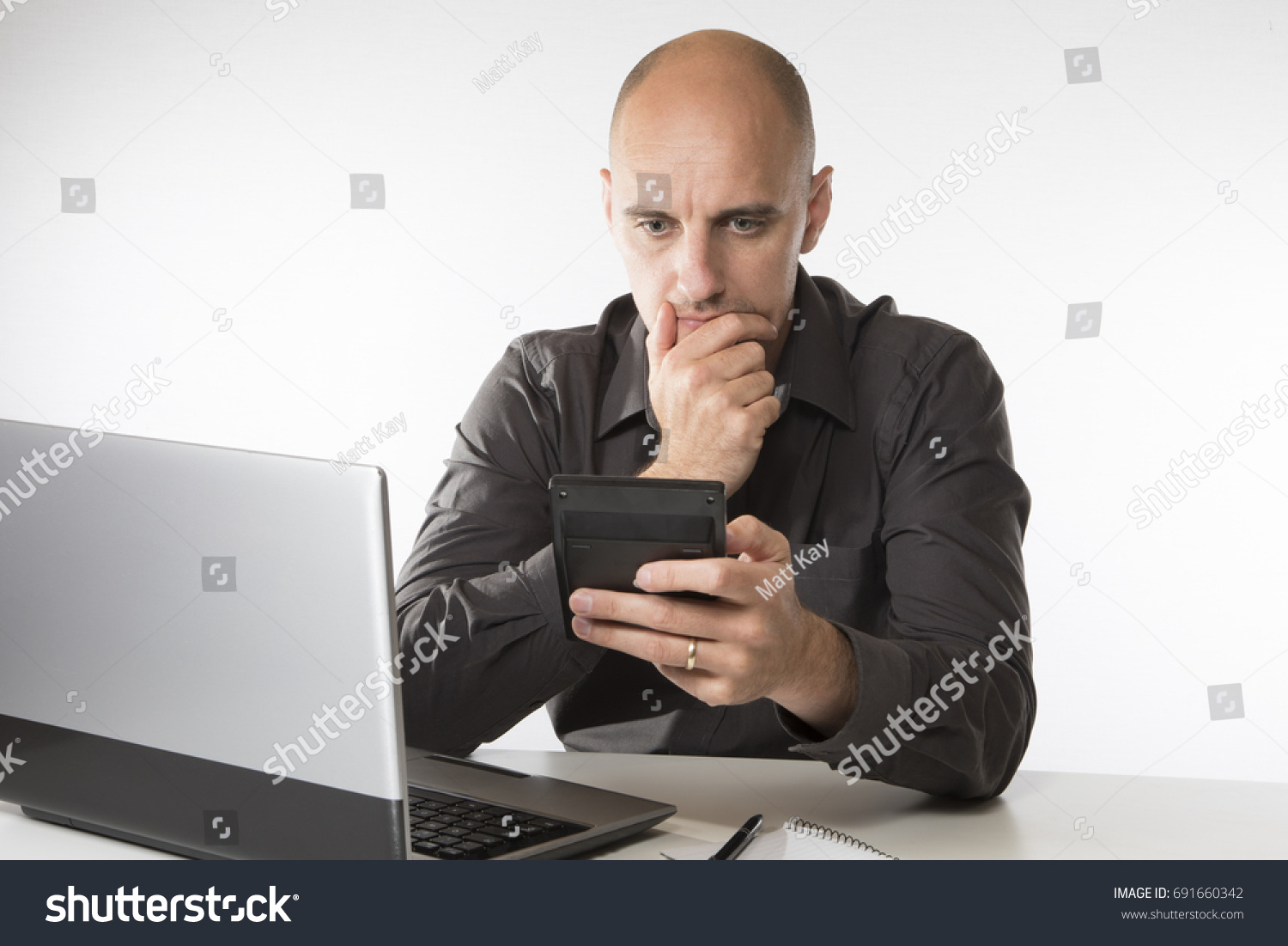 Worried man staring at a handheld calculator as he sits at his desk working on a laptop computer #691660342