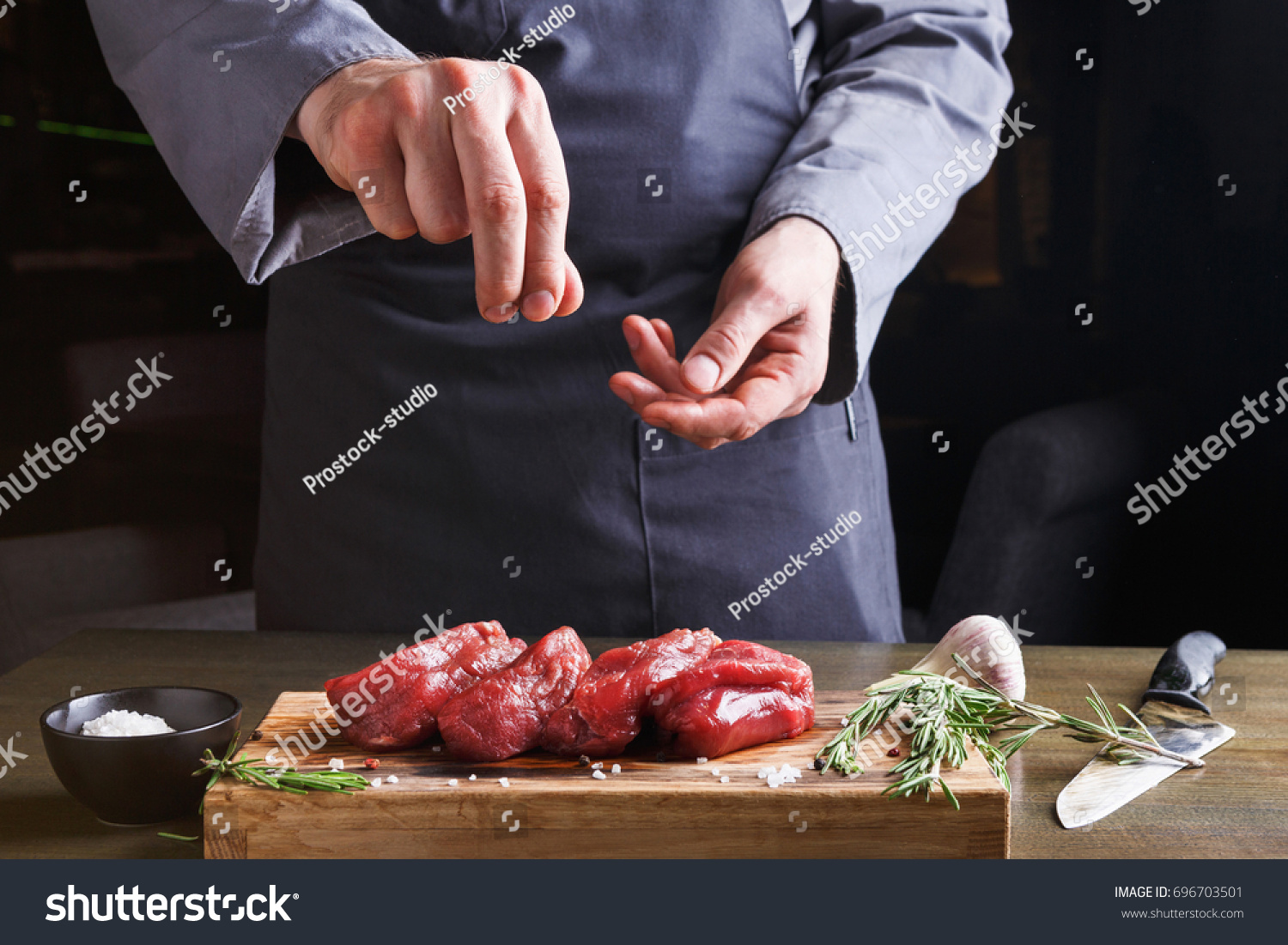 Man sprinkles filet mignon steaks with pepper salt. Chef working at open restaurant kitchen. Fresh meat, garlic and rosemary on wooden board. Modern restaurant cuisine backgroung #696703501