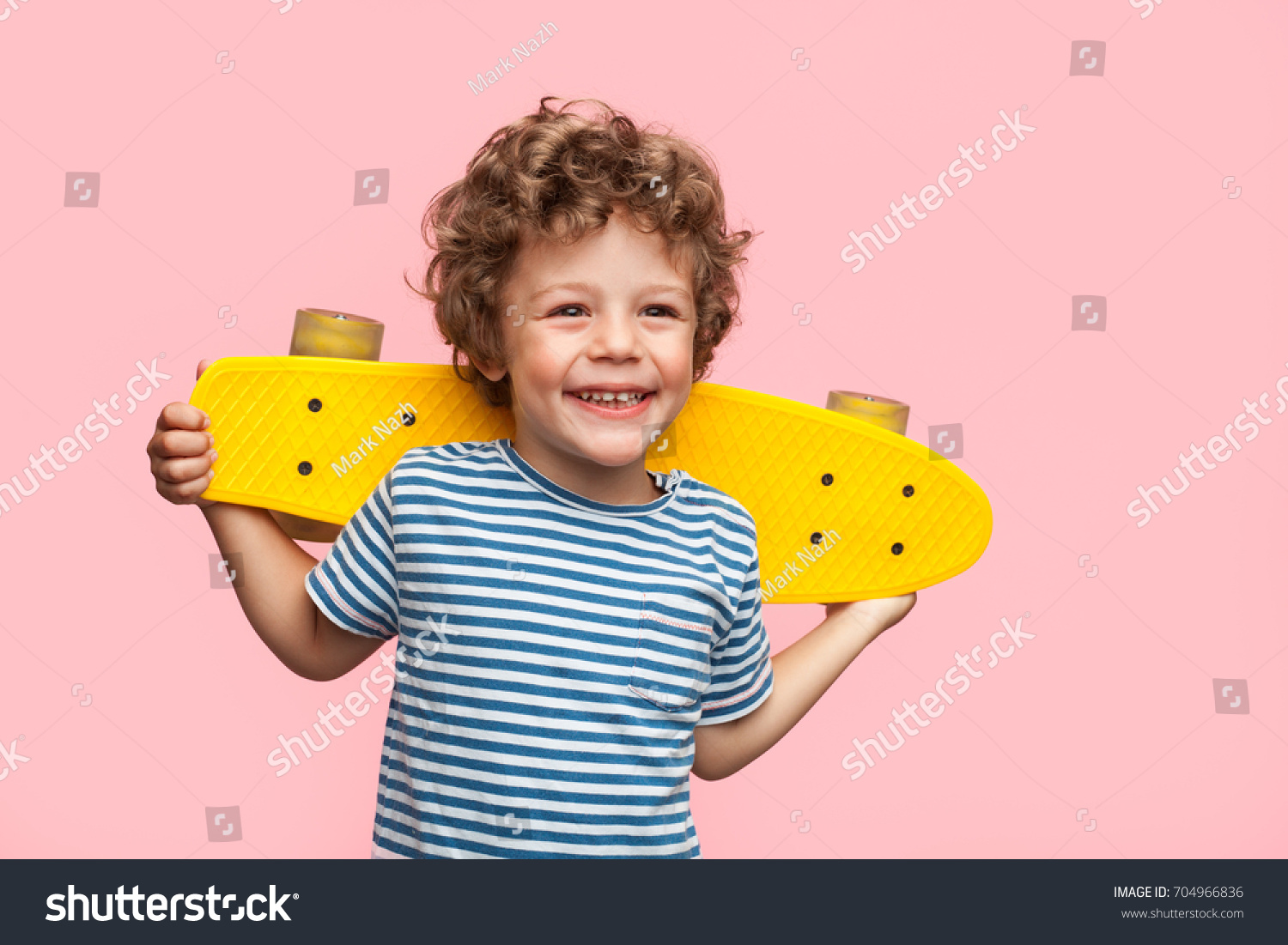 Little curly boy holding yellow longboard and looking away on pink background.  #704966836