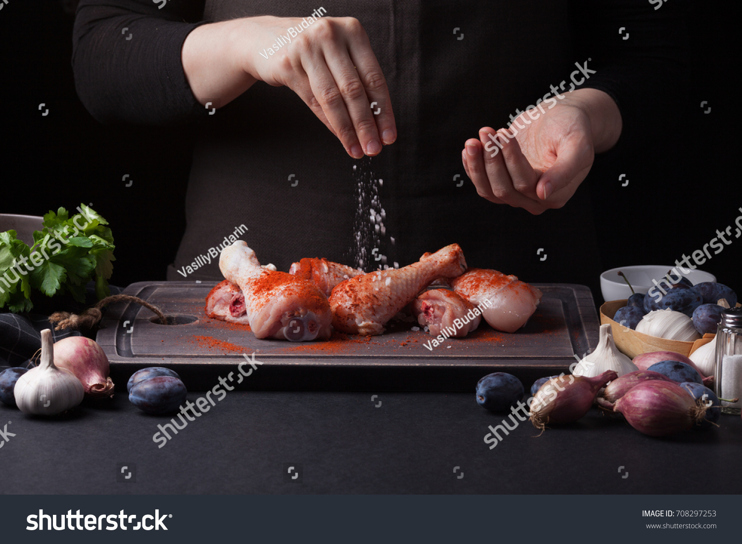 A female chef sprinkles fresh raw chicken drumsticks on a dark background with sea salt. Nearby lie the ingredients for cooking: shallots, blue plums, garlic pepper, salt and parsley. #708297253