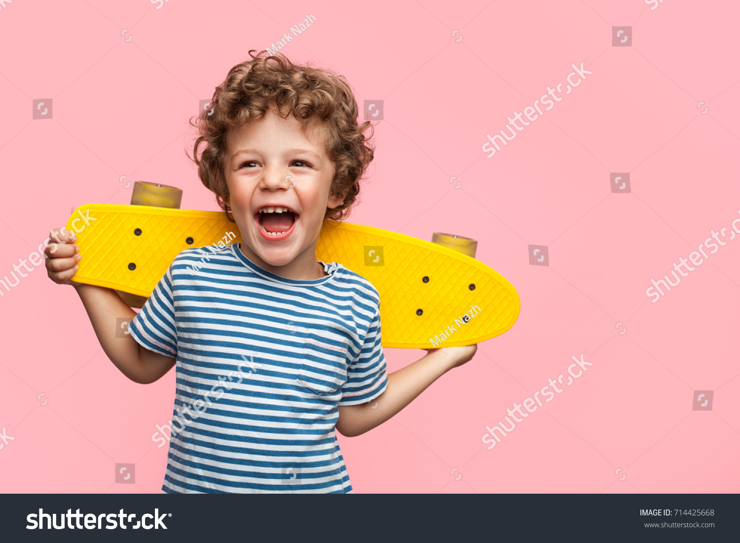 Charming curly boy holding yellow longboard and looking away on pink background.  #714425668