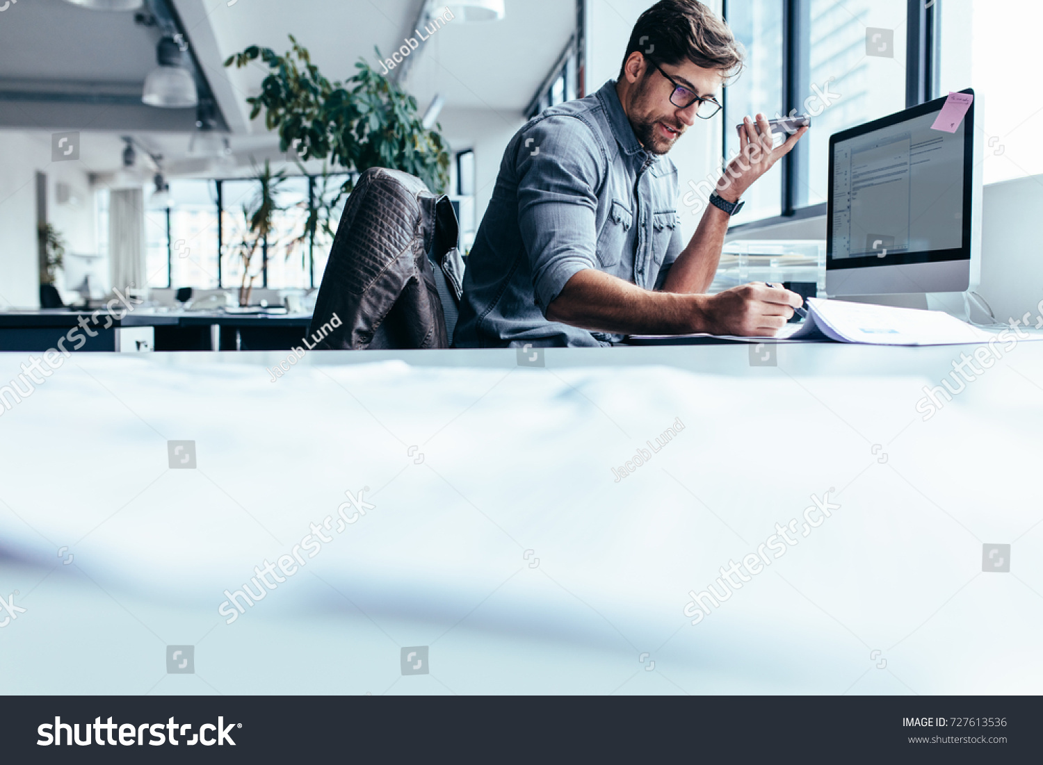Businessman talking on mobile phone and looking at documents. Young male executive working in office. #727613536