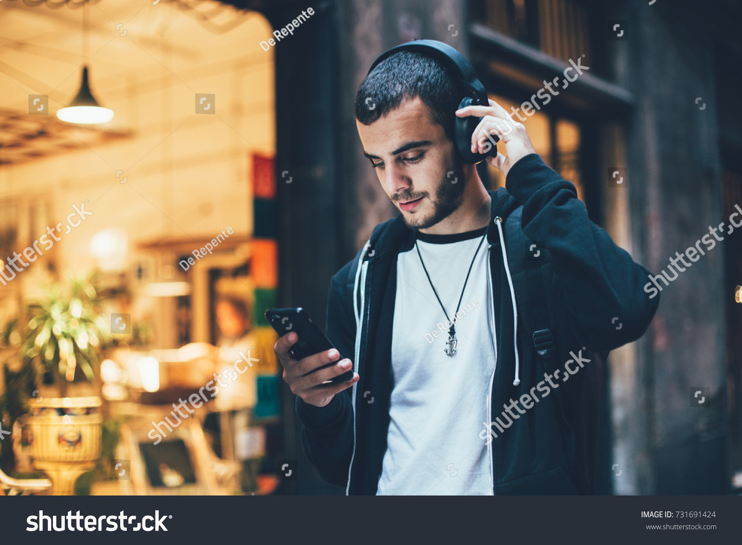 Hispanic young attractive man stands in dark street in front of shop, changes songs and tracks on smartphone, listens to music in wireless headphones. Hipster with slight beard #731691424