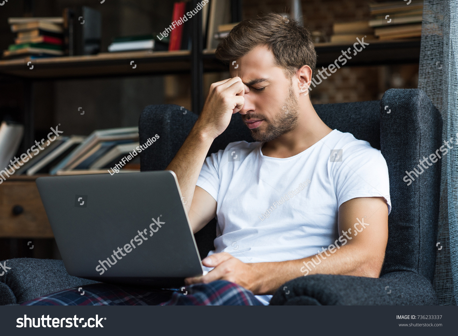 Young man in home clothes sitting in armchair with laptop and rubbing his nosebridge #736233337