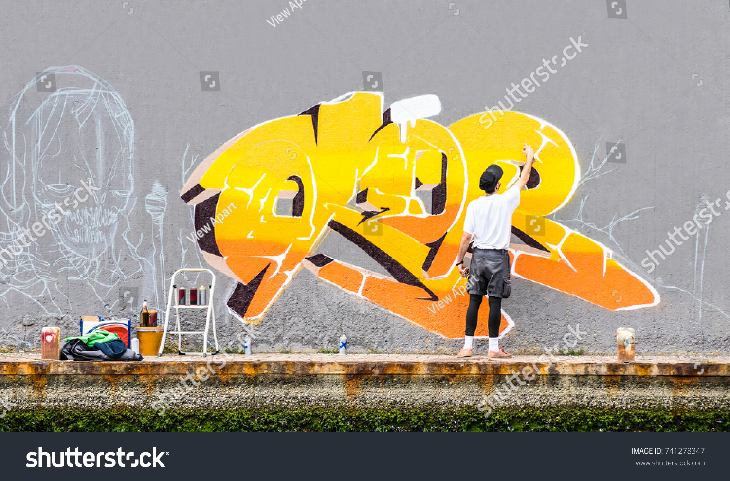Street artist painting colored graffiti on public space wall - Modern art concept of urban guy performing and preparing live murales paint with yellow aerosol color spray - Cloudy afternoon filter #741278347