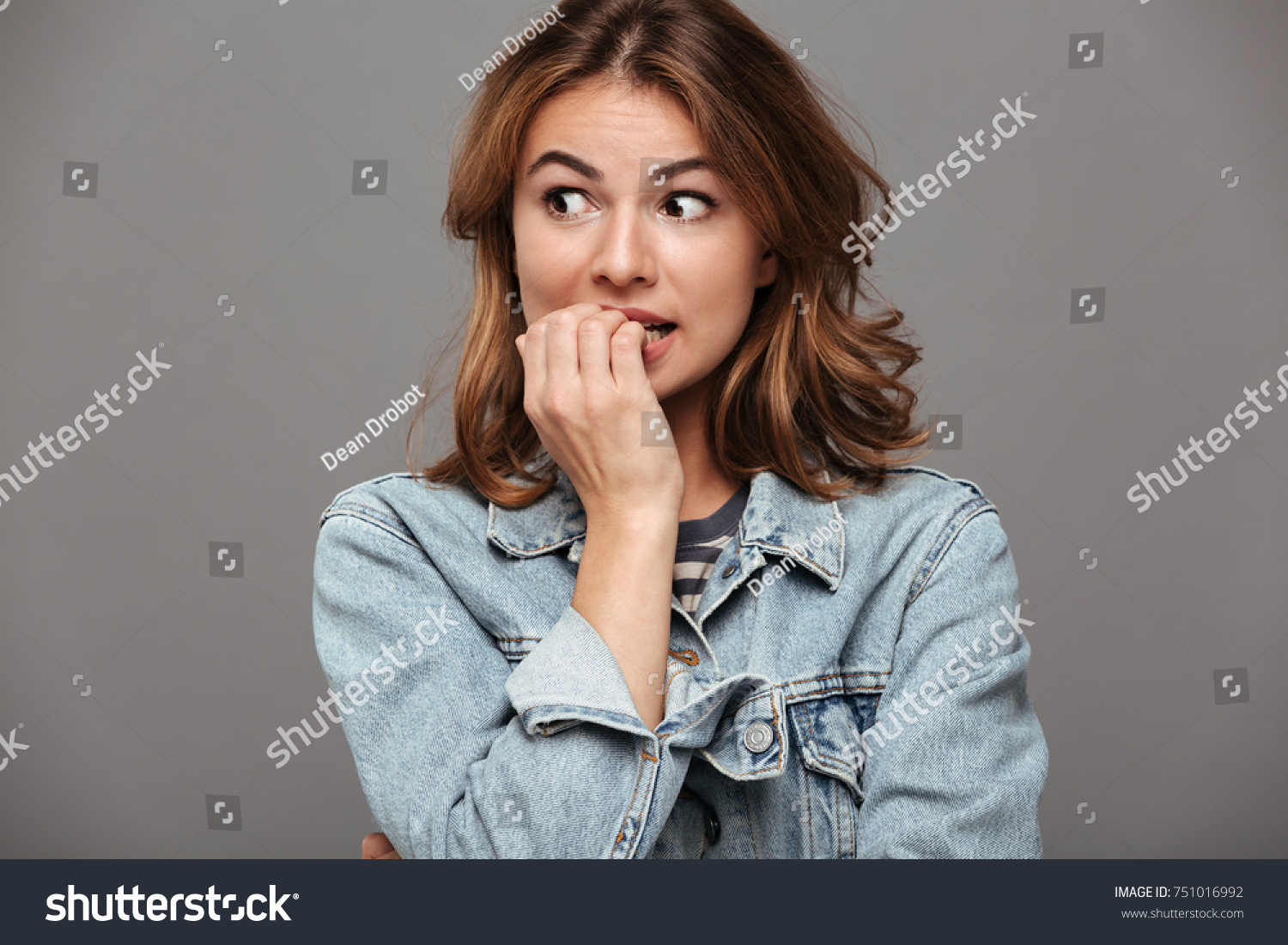 Close up portrait of a worried teenage girl in denim jacket biting her nails and looking away isolated over gray background #751016992