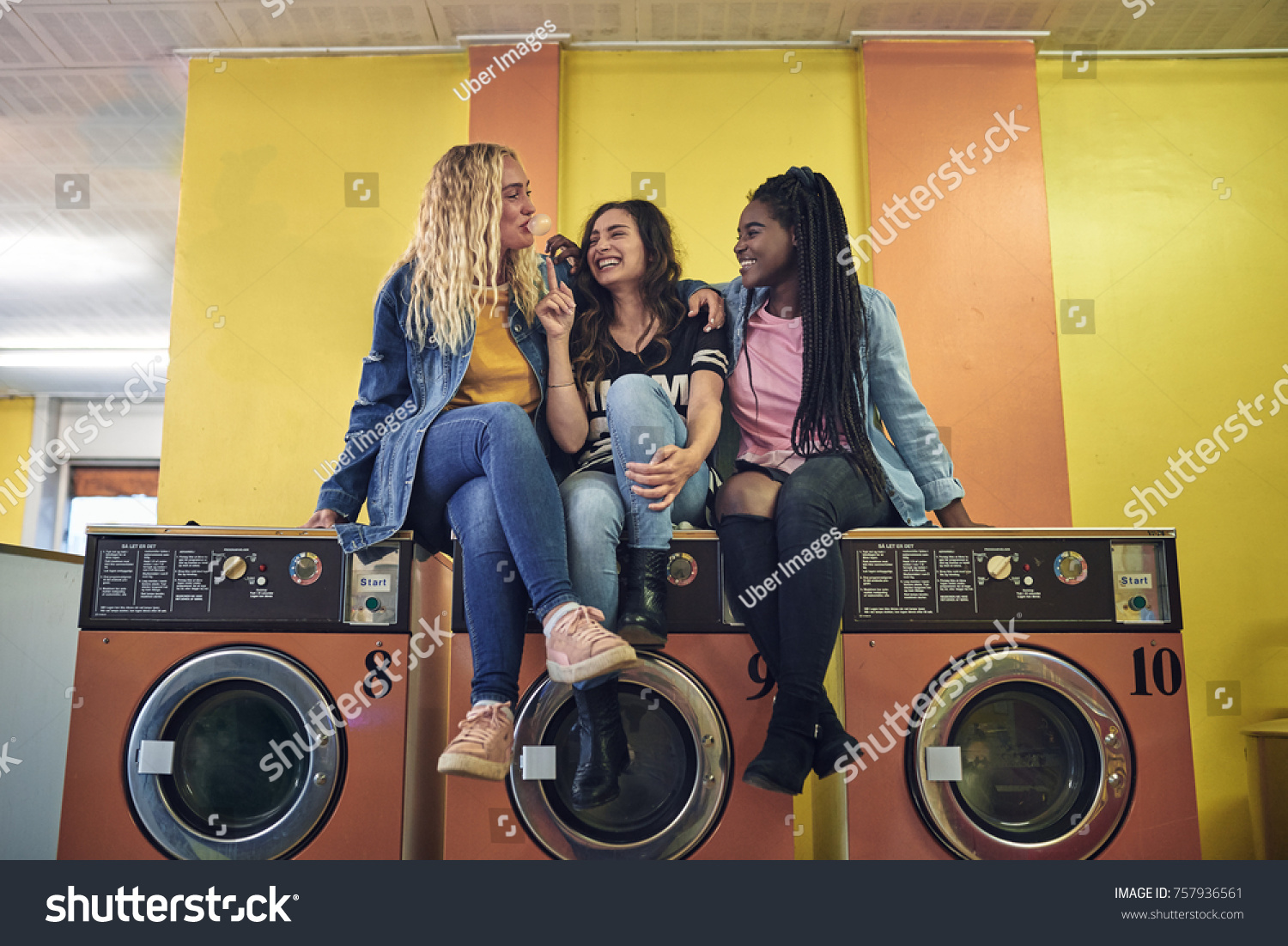 Group of young girlfriends sitting on washing machines in a laundromat together laughing and blowing bubbles with chewing gum #757936561