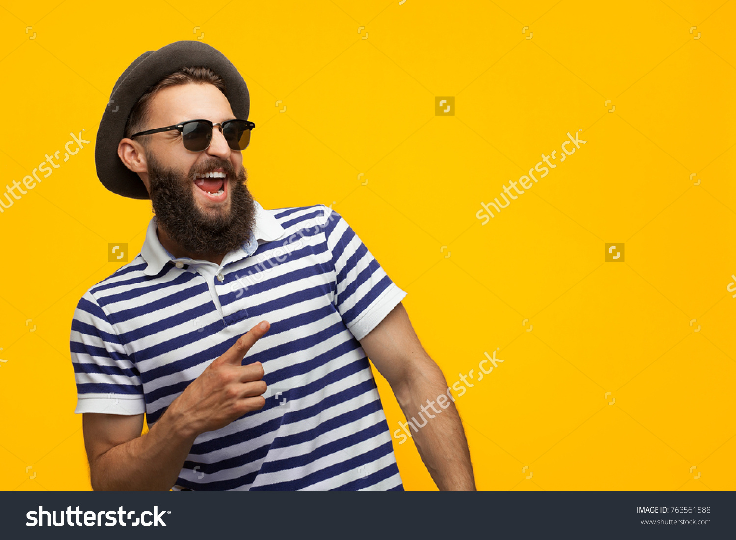 Young bearded hipster in cap and sunglasses pointing happily away on orange background.  #763561588