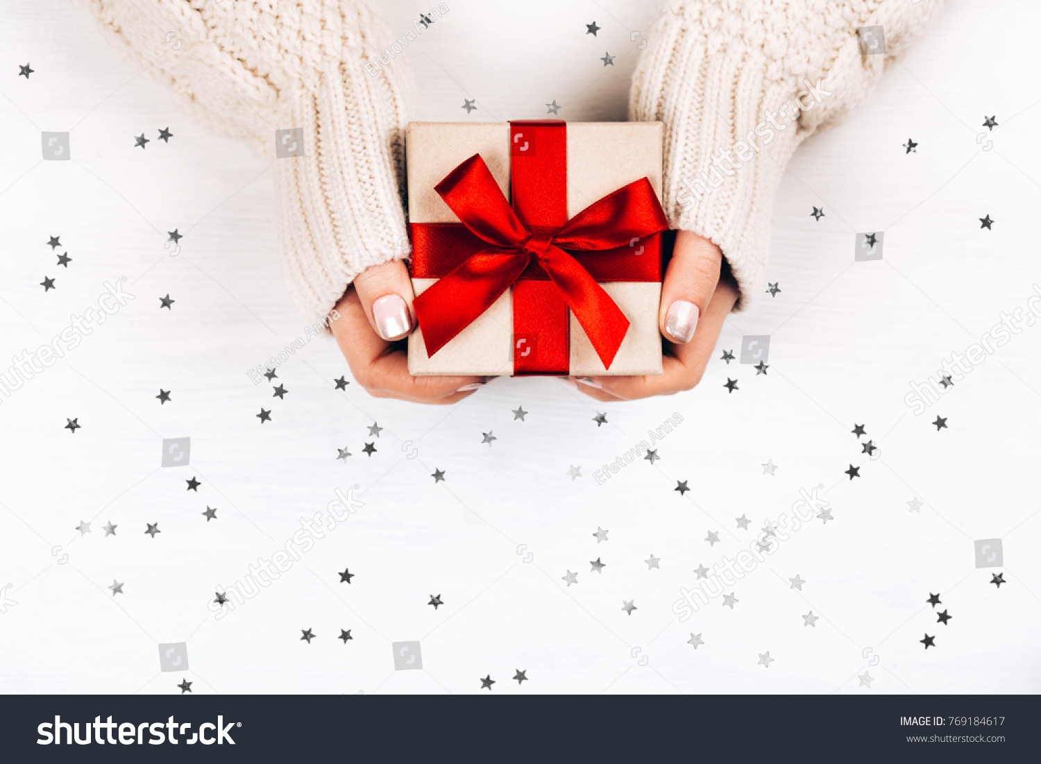 Female hands holding present with red bow on white rustic background with silver sparkles. Festive backdrop for holidays: Birthday, Valentines day, Christmas, New Year. Flat lay style #769184617