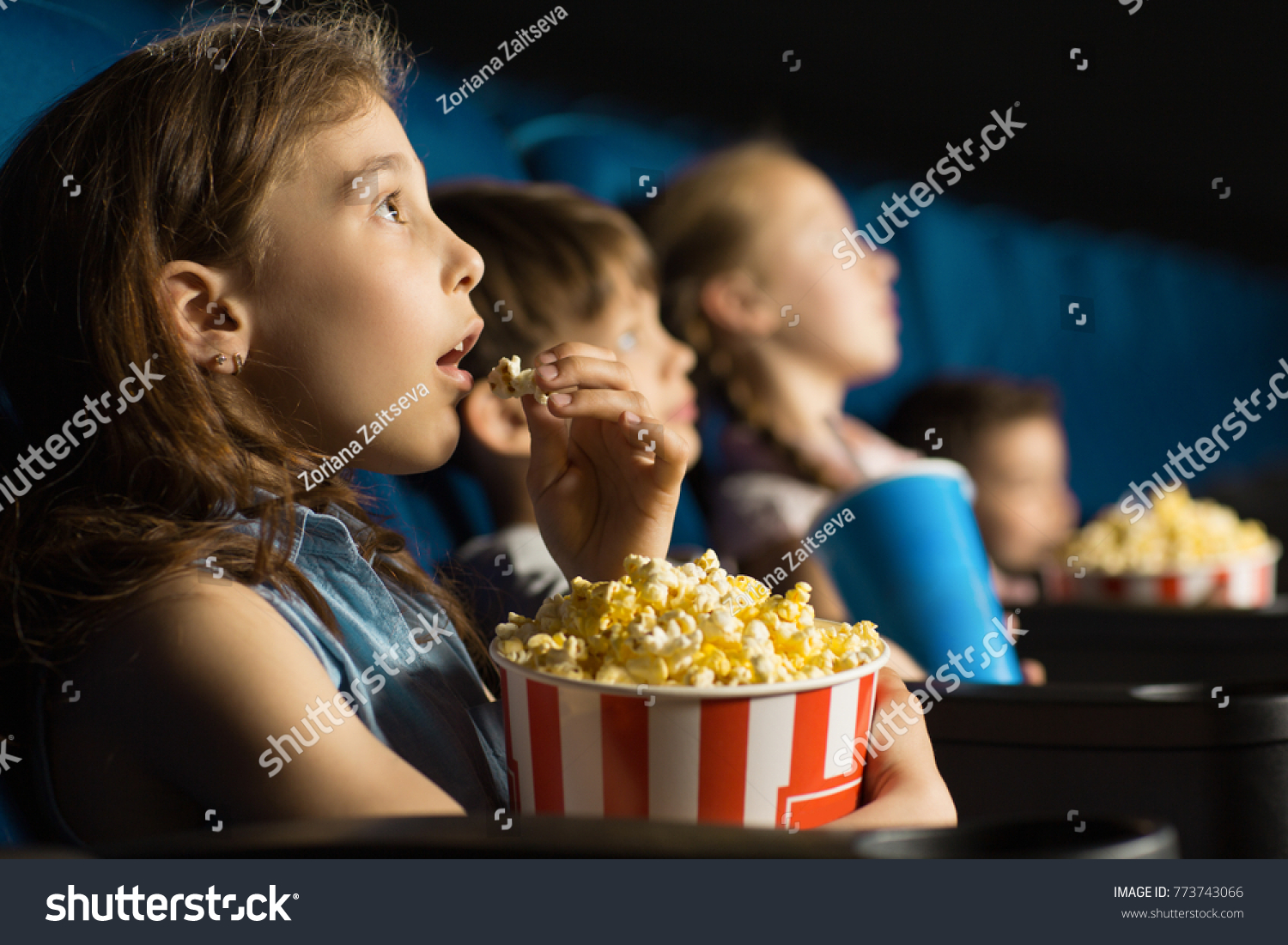 Beautiful little girl looking fascinated eating popcorn watching a movie at the local movie theatre snack bucket junk food tasty childhood entertaining entertained emotions kids concept #773743066