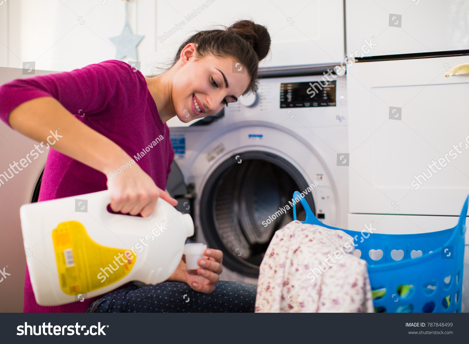 Portrait of the young smiling woman who sitting near washing machine in the room and pouring rinses into the lid #787848499