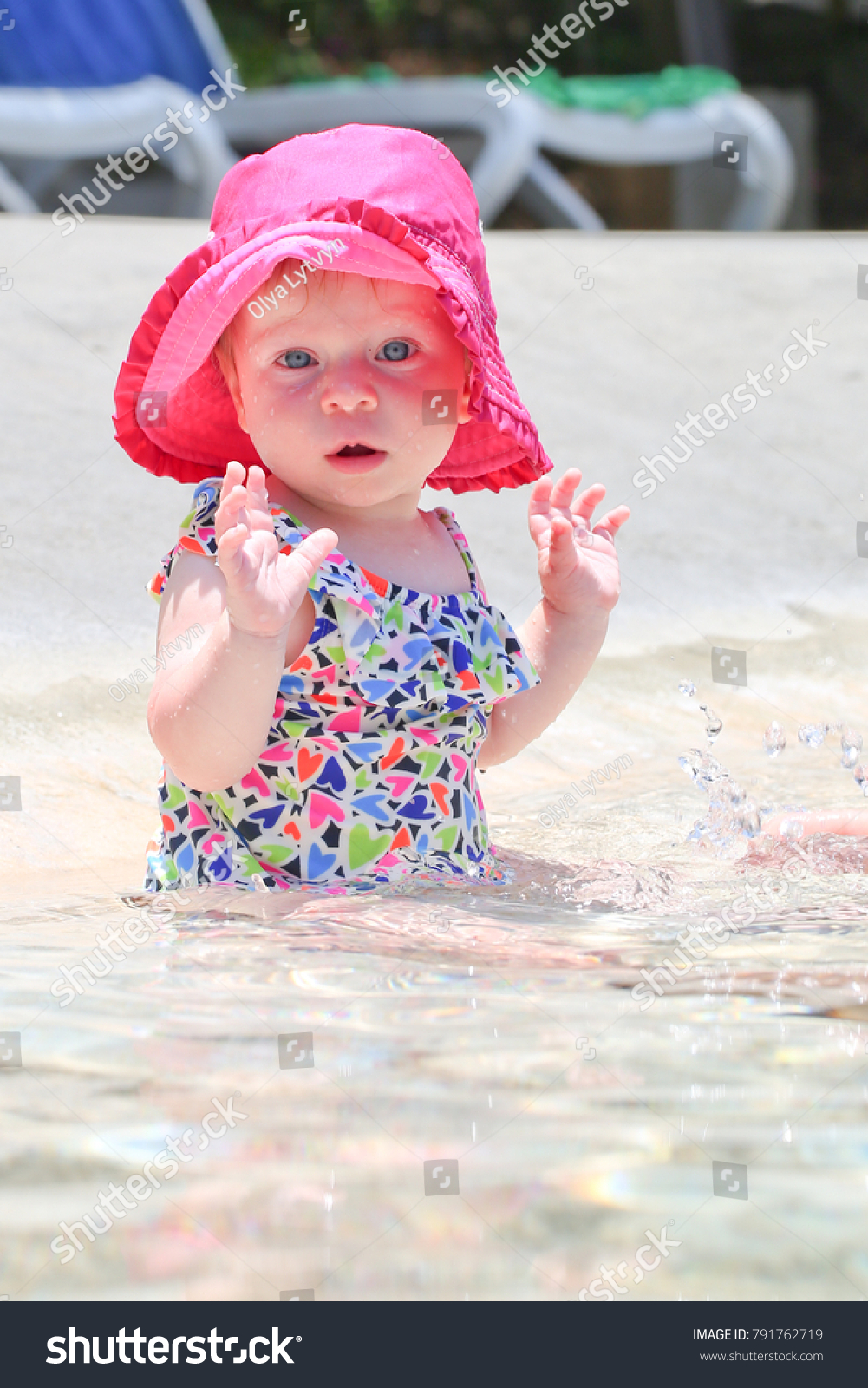 Cute adorable eight month old infant girl in pink hat and colorful swimsuit seat at the