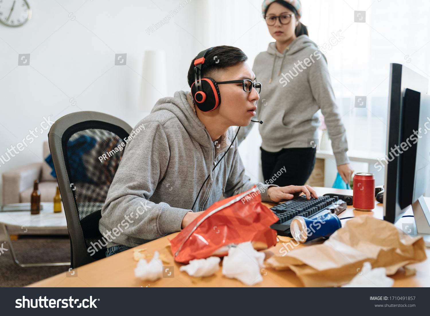 ボーイフレンドのテクノロジー中毒に悩まされた疑い深い彼女 家の散らかった部屋でコンピューター オンライン ゲームをしている間に 男性をじろじろ見て歩き続ける怒りに満ちた若いアジア人女性にボケ の写真素材 今すぐ編集