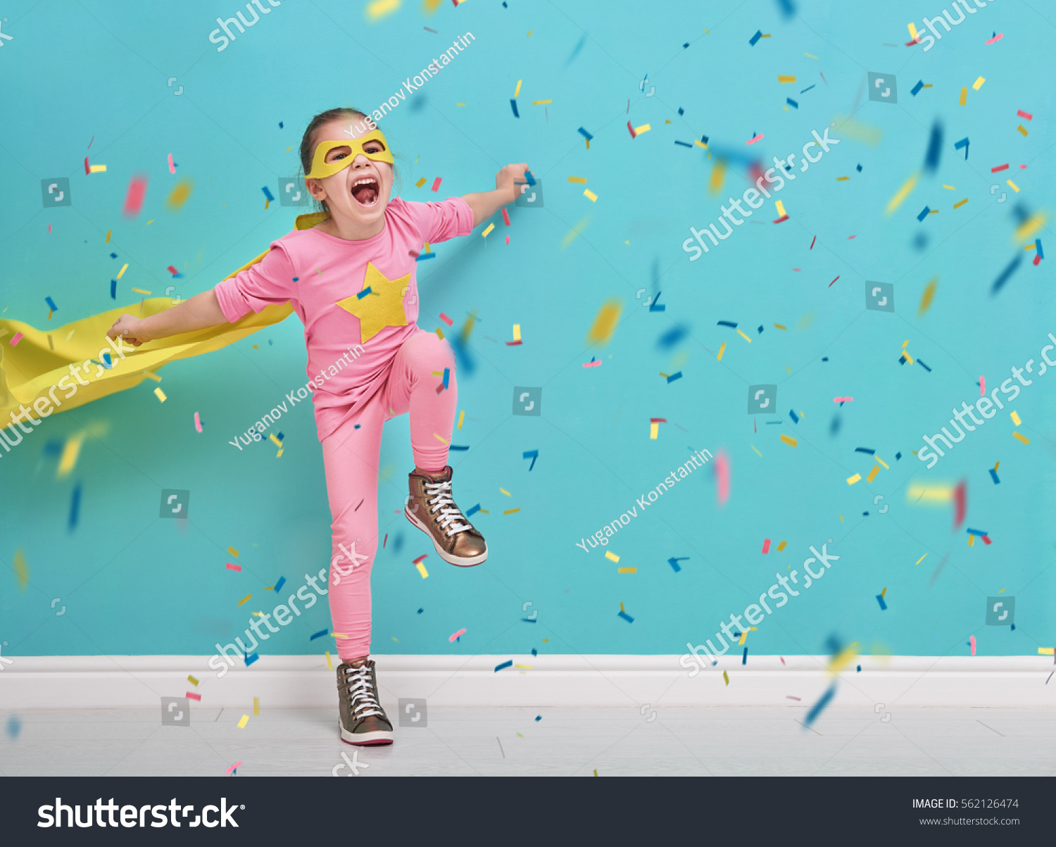 Little child plays superhero. Kid on the background of bright blue wall. Girl is throwing confetti and jumping. Yellow, pink and  turquoise colors.