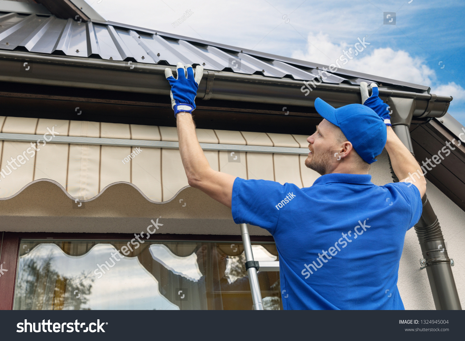 A man is standing on a ladder fixing a gutter on the roof of a house.