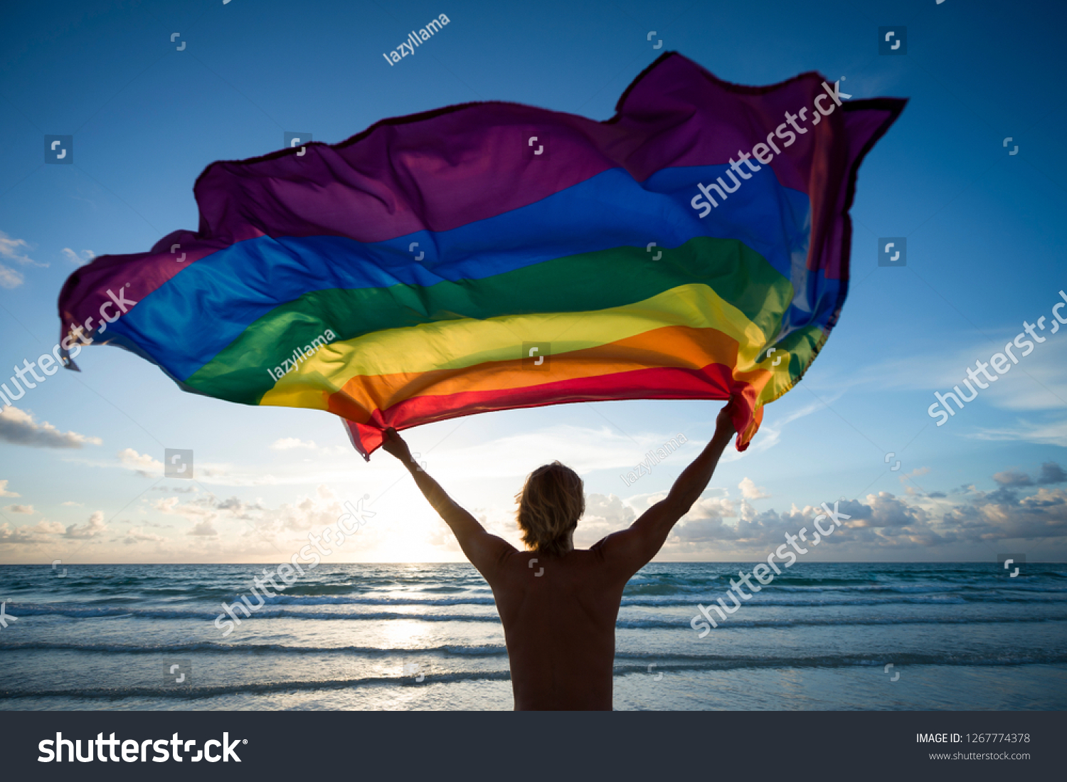 空の海岸で日の出にひらめく陽光に照らされる ゲイのプライドの虹の旗を持つ男性のシルエット の写真素材 今すぐ編集