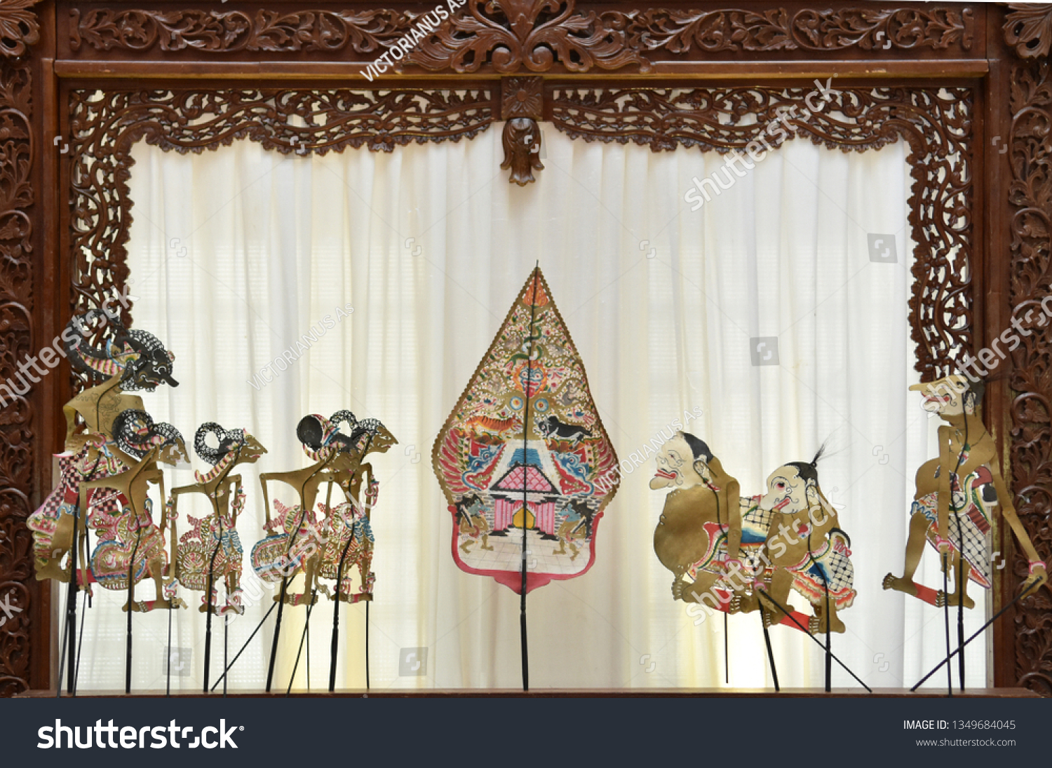  The image shows a collection of intricately designed wayang kulit (leather shadow puppets), depicting various mythological figures from the Hindu epic Mahabharata and Ramayana, displayed against a white backdrop with a wooden frame.