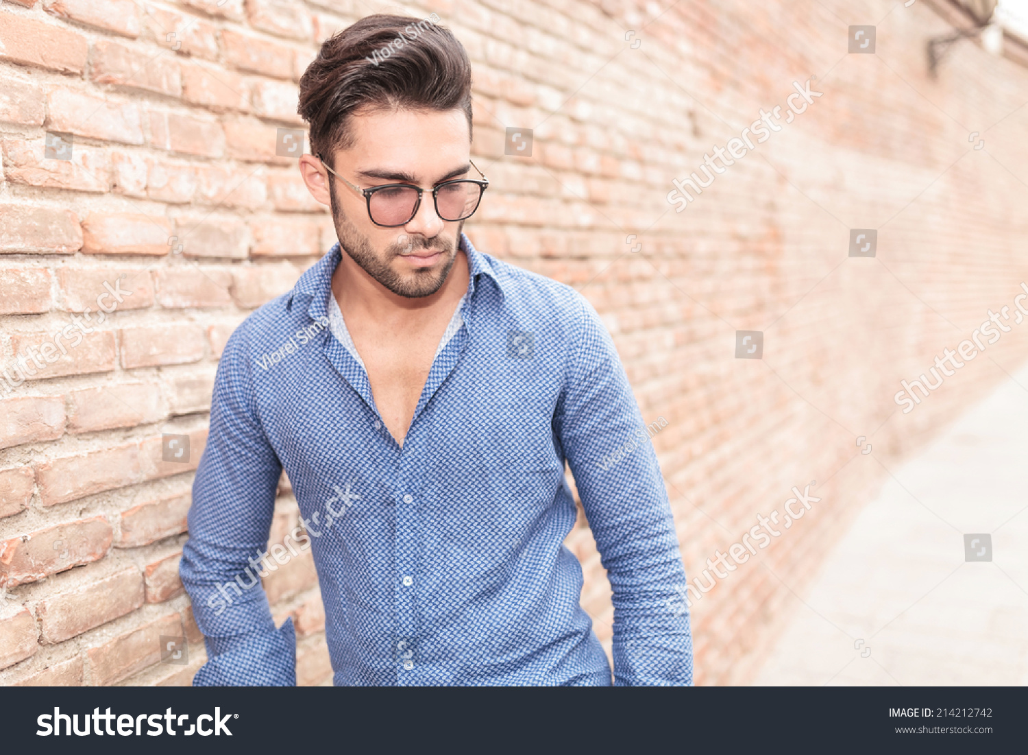 Young Casual Man With Glasses Looks Down While Standing Near Brick Wall ...