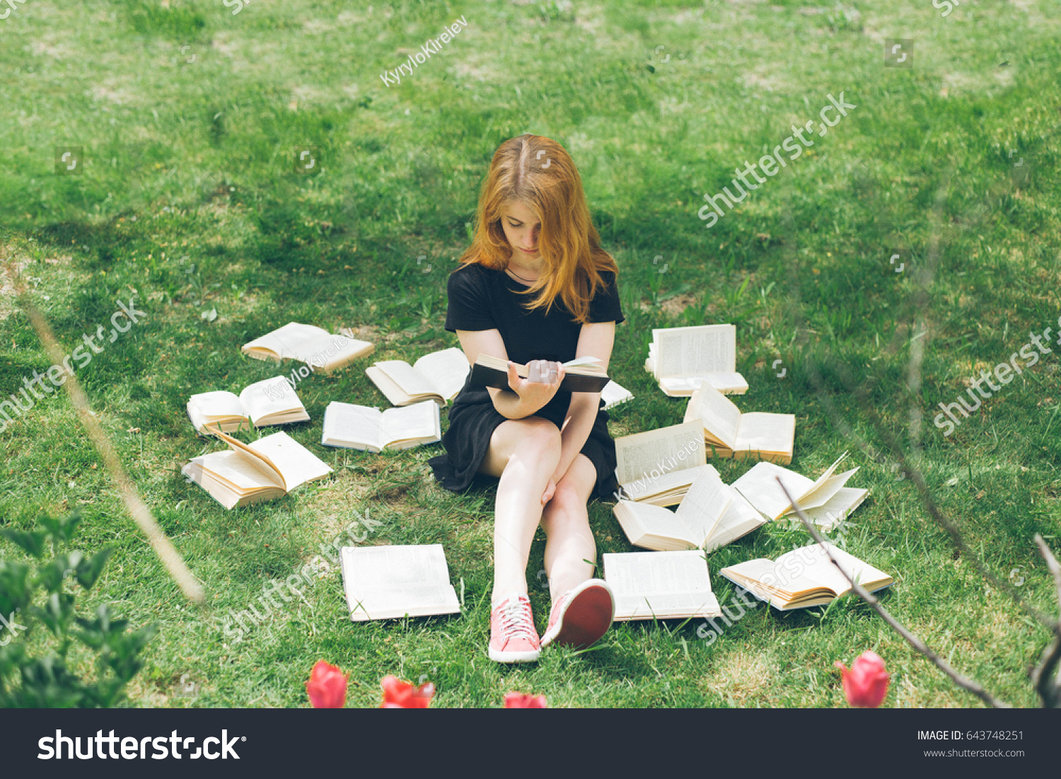 Young girl reading a book while lying in the grass. A girl sits among the many books in the garden. Many books on the grass in the summer garden