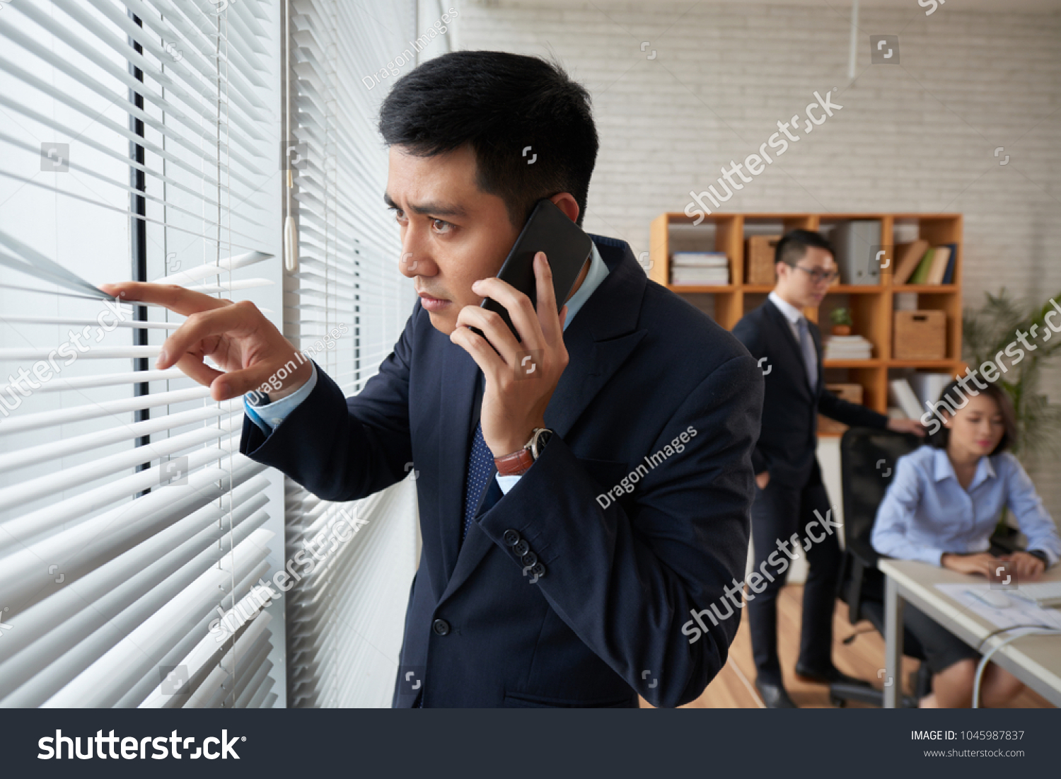 Young Vietnamese Businessman Looking Through Blinds On Office Window When Calling On The Phone